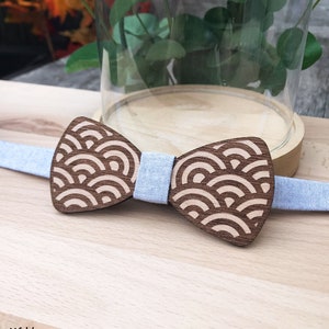 May include: A wooden bow tie with a light blue denim strap. The bow tie features a geometric pattern of overlapping arcs in a light brown colour.
