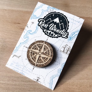 May include: A wooden compass pin with a detailed design, featuring a compass rose and a star. The pin is displayed on a white card with a blue and white map design. The card also features a black and white logo with the text "Ron Woody".