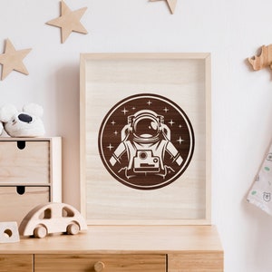 Könnte beinhalten: Ein gerahmter Druck eines braunen Astronauten in einem Raumanzug mit einem weißen Helm, umgeben von weißen Sternen in einem Kreis auf einem hellbraunen Hintergrund.