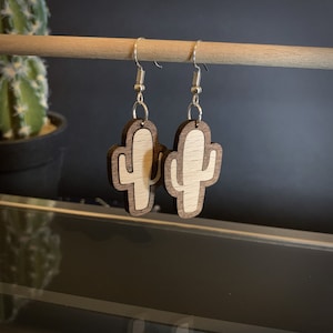 May include: A pair of wooden cactus earrings. Each earring features a light-colored cactus shape set within a darker wood frame, suspended from silver-colored ear wires. The earrings are displayed against a neutral background.
