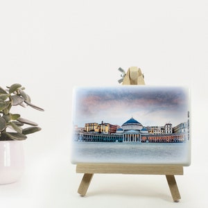 May include: A white ceramic tile with a printed image of a cityscape. The tile is on a small wooden easel. The cityscape includes a large domed building and other buildings in the background. The sky is a soft pink and orange.