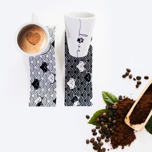 Puede incluir: Dos tazas de cer&aacute;mica blanca con dise&ntilde;os de gatos negros y la palabra "Love" escrita en una de las tazas. Las tazas est&aacute;n sobre posavasos con patrones en blanco y negro. Granos de caf&eacute; y caf&eacute; molido est&aacute;n esparcidos alrededor de las tazas.