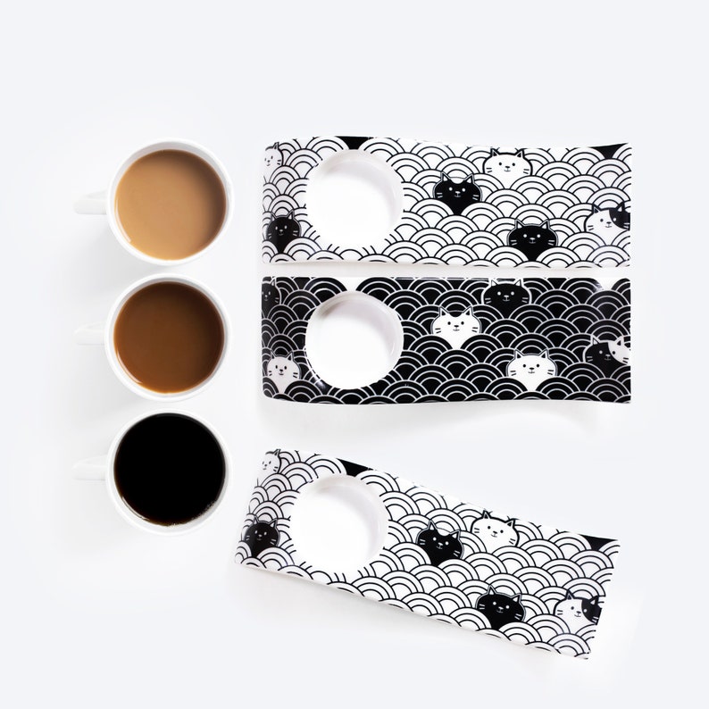 Puede incluir: Tres tazas de cer&aacute;mica blanca con bordes y asas negras, llenas de caf&eacute; o t&eacute;, se muestran sobre una superficie blanca. Tambi&eacute;n se muestran dos posavasos de cer&aacute;mica en blanco y negro con un patr&oacute;n repetitivo de gatos blancos y negros y olas.