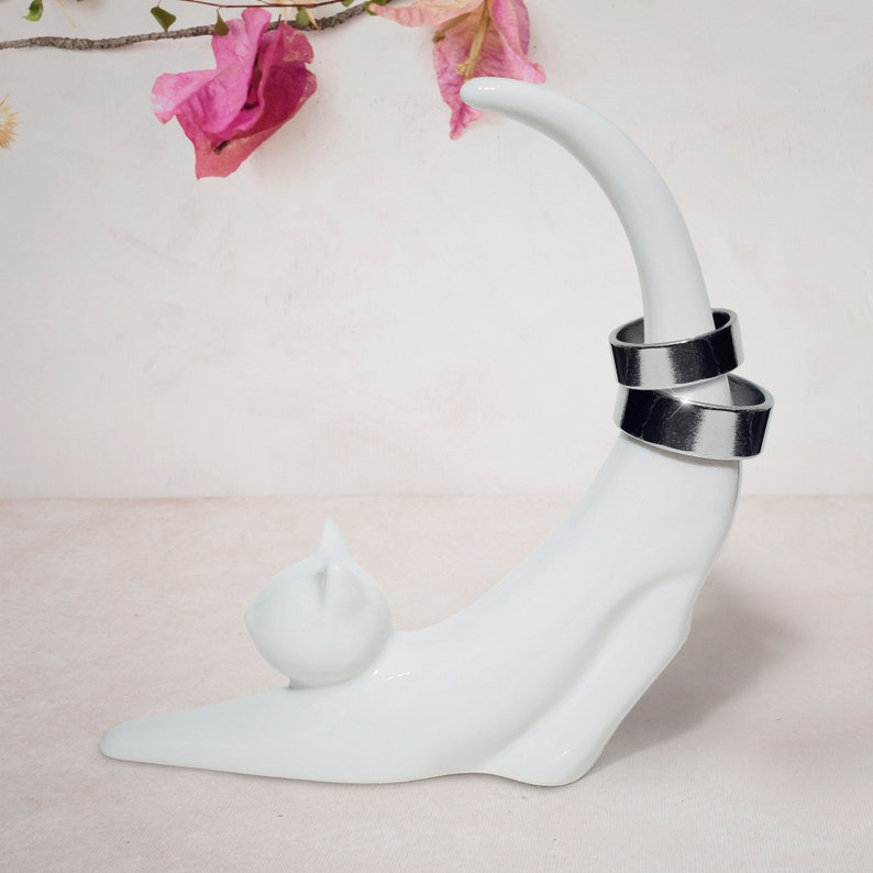 May include: White ceramic ring holder shaped like a cat curled up. The cat's tail is curved upwards and holds two silver rings.