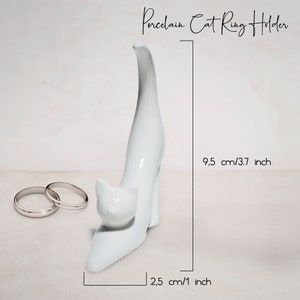 May include: White porcelain cat ring holder with a raised tail. The cat is standing on its hind legs with its front paws resting on the ground. The holder measures 9.5 inches tall and 2.5 inches wide.