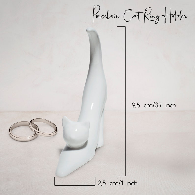 May include: White porcelain cat ring holder with a raised tail. The cat is standing on its hind legs with its front paws resting on the ground. The holder measures 9.5 inches tall and 2.5 inches wide.