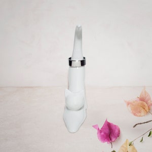May include: White ceramic cat ring holder with a silver ring on the top. The cat is sitting up with its tail curled around its body. The background is a white surface with pink flowers.