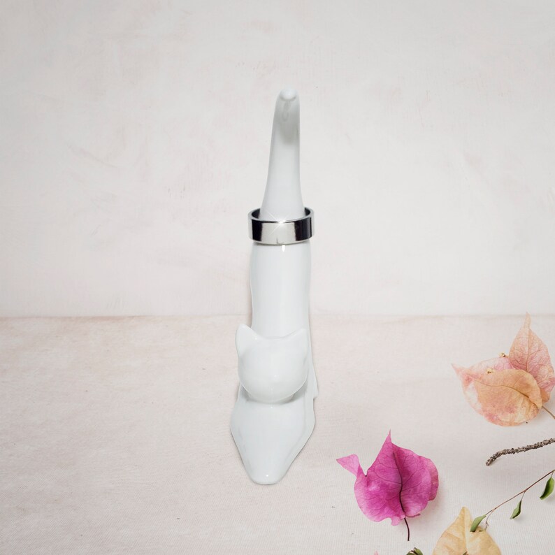 May include: White ceramic cat ring holder with a silver ring on the top. The cat is sitting up with its tail curled around its body. The background is a white surface with pink flowers.