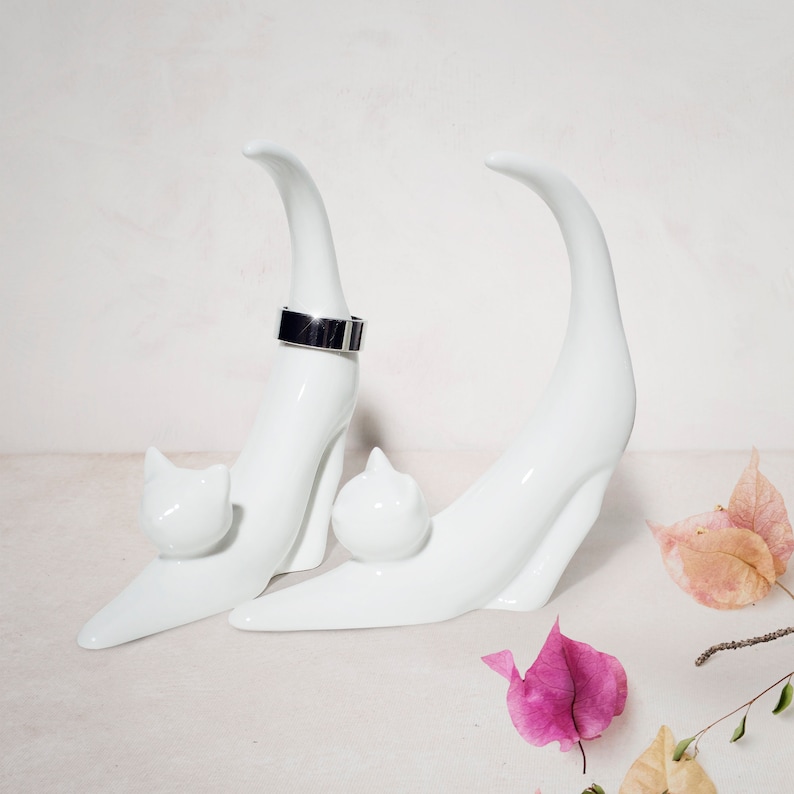 May include: White ceramic cat ring holder with a silver ring on the top. The cat is in a sitting position with its tail curled around its body. The ring holder is on a white surface.