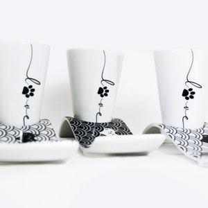 Puede incluir: Tres tazas de espresso de cer&aacute;mica blanca con huellas de patas negras y la palabra "love" escrita en ellas. Cada taza est&aacute; sobre un platillo blanco y negro con un patr&oacute;n de ondas.