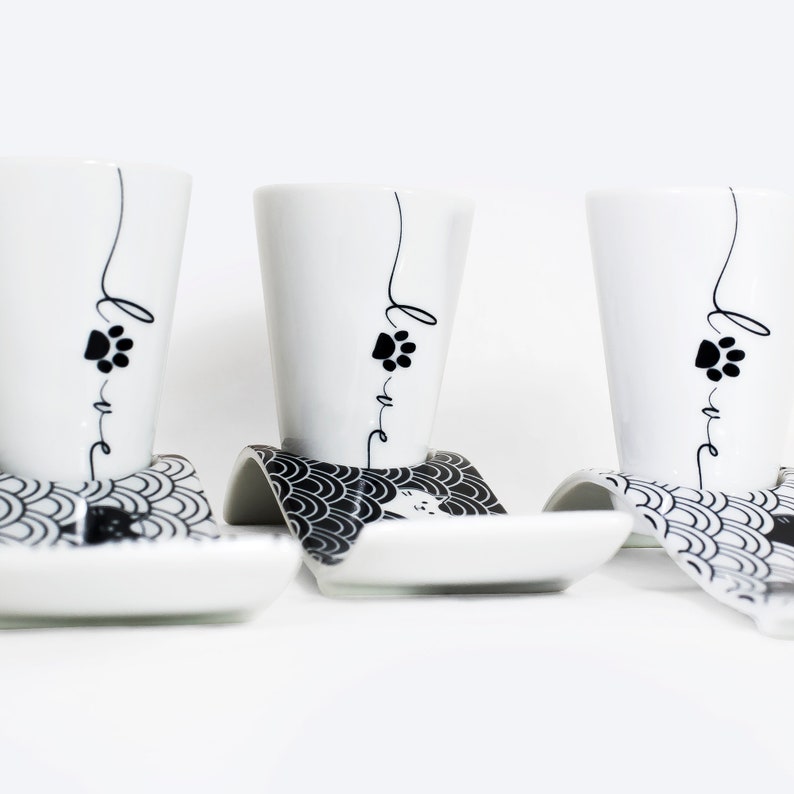 Puede incluir: Tres tazas de espresso de cer&aacute;mica blanca con huellas de patas negras y la palabra "love" escrita en ellas. Cada taza est&aacute; sobre un platillo blanco y negro con un patr&oacute;n de ondas.