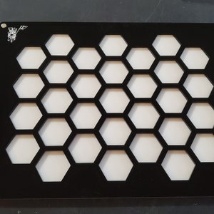 Pode incluir: Painel de exibição de acrílico preto com um padrão de favo de mel cortado. O painel tem 25 espaços hexagonais para exibir itens pequenos.
