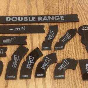 May include: Black and white printed plastic pieces for a board game. The pieces include a double range, small template, large template, slide, veer, swerve, gentle, turn, hard, hairpin, long, medium, and short.