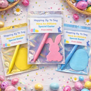 May include: Three Easter-themed gift bags with pens and egg or bunny-shaped cutouts. Each bag has a label that reads "Hopping By To Say Have An EGGstra Special Easter From Ashley". The pens are pink and blue.
