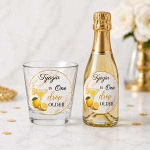 May include: A clear shot glass and a small champagne bottle with gold foil tops. Both have a circular label with the text "Tyazia is One drop OLDER" and a lemon cocktail illustration. The background is a marble pattern.