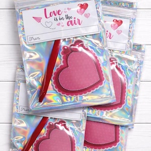 May include: Several holographic bags with a pink heart-shaped tag and a red pen inside. The tag reads "Love is in the air" with a heart and airplane graphic. The bags are sealed and ready for gifting.