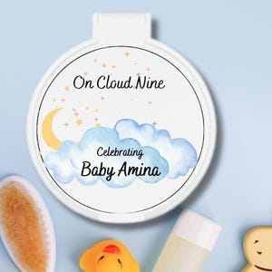 May include: A white circular baby shower favor with the text "On Cloud Nine Celebrating Baby Amina" surrounded by a watercolor design of clouds, a crescent moon, and stars. The image also includes a baby brush, a rubber duck, a bottle, and a bunny-shaped toy.