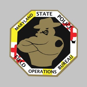May include: An octagonal Maryland State Police Field Operations Bureau emblem. The emblem features a brown figure with crossed arms wearing a hat, surrounded by text in yellow and white. Red and white accents are present.
