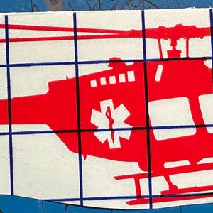 Może przedstawiać: Czerwona sylwetka helikoptera z białym symbolem gwiazdy życia po bokach. Helikopter znajduje się na białym tle.