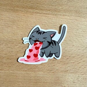 May include: A sticker featuring a gray cartoon cat with black stripes, vomiting a stream of red hearts. The cat has a happy expression, and the vomit is pink. The sticker has a white border.