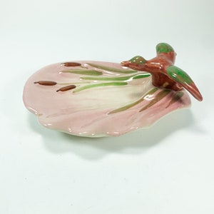 Puede incluir: Un plato de cerámica rosa con forma de nenúfar con una figurita de pájaro verde y marrón en la parte superior. El pájaro está volando fuera del plato.