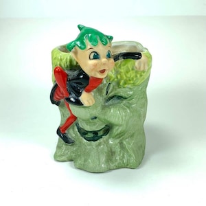 May include: A green ceramic planter shaped like a tree stump with a green-haired elf peeking out from the side. The elf is wearing a black and red outfit and has blue eyes.