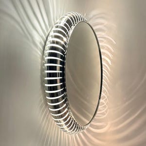 Op de afbeelding: Een ronde wandspiegel met een metalen, geribbelde lijst met geïntegreerde verlichting. De lijst bestaat uit afwisselende zilverkleurige segmenten en verlichte witte banden die een radiaal lichtpatroon op de omliggende muur werpen.