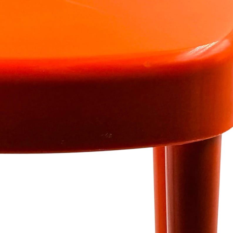 May include: Close-up of a bright orange chair with a rounded seat and legs. The chair's color transitions from a vibrant orange on top to a deep red at the base, creating a gradient effect. The legs are cylindrical and the background is white.