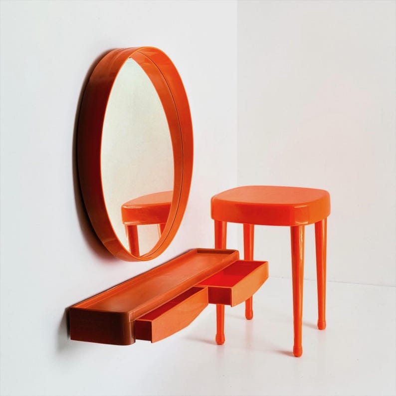 May include: An orange, modern vanity set with an oval mirror, a shelf with a drawer, and a small stool. The mirror is mounted on the wall, and the shelf is attached below it. The stool has four legs.