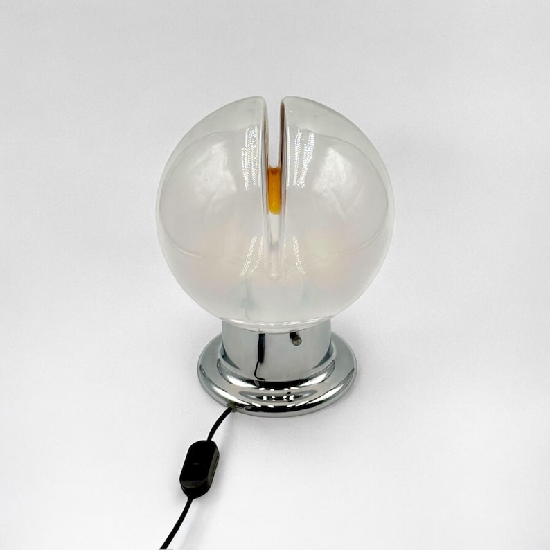 May include: A white glass table lamp with a chrome base and a split globe design. The lamp has a black cord and switch.