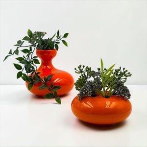 Könnte beinhalten: Zwei glänzende orangefarbene Keramikvasen mit glatten, abgerundeten Formen. Eine Vase hat einen hohen, schmalen Hals, während die andere eine niedrige, breite Schale mit mehreren Öffnungen ist. Beide enthalten künstliches grünes Blattwerk mit grauen Beeren.