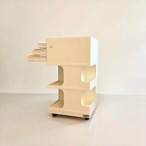 Può includere: Un mobiletto contenitore mobile color crema con una sezione superiore dotata di tre piccoli cassetti estraibili e una serratura a chiave. La base è composta da due ripiani aperti sostenuti da pilastri curvi, montati su piccole ruote nere.
