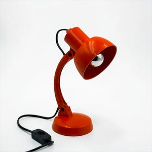 May include: A bright orange desk lamp with a curved adjustable arm and a round base. The lamp features a glossy finish, a wide conical shade, and a silver-toned bulb. A black power cord with an integrated switch extends from the base.