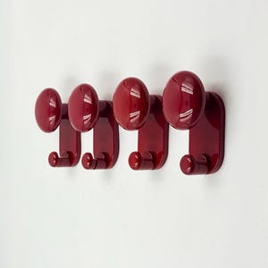 Puede incluir: Cuatro perchas de pared rojas brillantes. Cada percha tiene un pomo redondeado y un gancho inferior para colgar objetos. Las perchas están montadas en una pared blanca, ofreciendo un aspecto limpio y moderno.