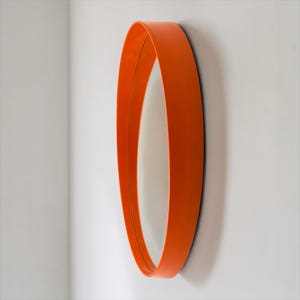 May include: An oval-shaped wall mirror with a bright orange frame. The mirror is mounted on a white wall, and the frame has a smooth, curved design. The mirror's reflective surface is visible within the frame.
