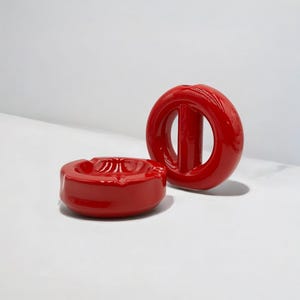 May include: A glossy red ceramic ashtray with a circular indentation and a matching glossy red circular object featuring a central vertical bar. Both items have a smooth, reflective finish and a vibrant, solid red colour throughout their surfaces.