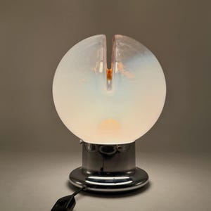 May include: A white glass table lamp with a chrome base. The lampshade is a sphere with a split in the top. The lamp is lit and casting a warm glow.