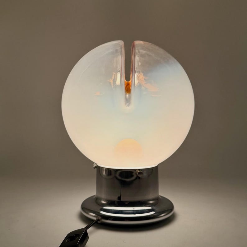 May include: A white glass table lamp with a chrome base. The lampshade is a sphere with a split in the top. The lamp is lit and casting a warm glow.