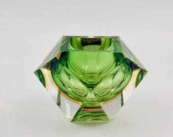May include: A faceted, decorative glass bowl with a geometric design. The bowl features a vibrant green interior and clear, angled exterior walls. Gold accents are visible between the green and clear glass, creating a striking contrast. The bowl is designed to catch and reflect light.