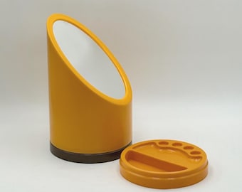 May include: A mustard yellow vanity mirror with a white interior and a matching round base with a small tray. The mirror has a cylindrical shape with a slanted top. The base is a separate piece.