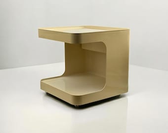 May include: A beige, modern side table with a unique C-shaped design. The table has a flat top and a lower shelf, connected by a curved side. It is made of a solid material and sits on small wheels.