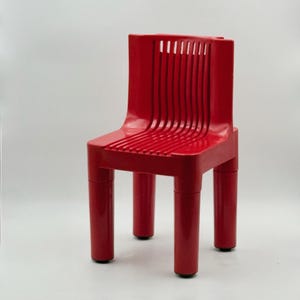 May include: A vibrant red chair with a unique design. The chair features a slatted seat and backrest, supported by four cylindrical legs. The backrest has vertical slits. The chair is made of a solid material and has a glossy finish.