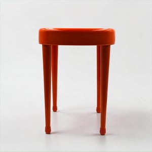 May include: A vibrant orange stool with a rounded seat and four slender, tapered legs. The legs have small, rounded feet. The stool is a solid color and appears to be made of plastic or a similar material. The design is modern and minimalist.