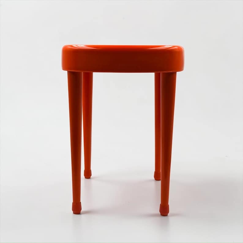 May include: A vibrant orange stool with a rounded seat and four slender, tapered legs. The legs have small, rounded feet. The stool is a solid color and appears to be made of plastic or a similar material. The design is modern and minimalist.