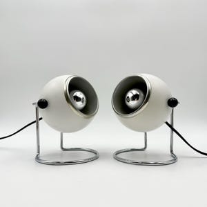 May include: Two white, spherical desk lamps with chrome accents and a black power cord. Each lamp has a silver-colored bulb and sits on a chrome stand with a black knob. The lamps are designed in a retro style.