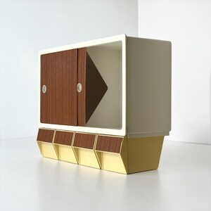 K&ouml;nnte beinhalten: Ein Retro-Stauraumschrank mit wei&szlig;em Rahmen, brauner Schiebet&uuml;r und gelbem Sockel. Der Schrank hat ein geometrisches Design mit vier kleinen Schubladen und einem dreieckigen Innenelement.