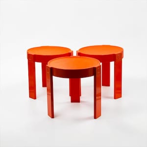 Könnte beinhalten: Drei leuchtend orangefarbene Hocker mit runder Sitzfläche und vier geraden Beinen. Jeder Hocker hat eine glatte, einheitliche Oberfläche und eine Konstruktion, bei der die Beine mit einem kreisförmigen Detail am Sitzrahmen befestigt sind.