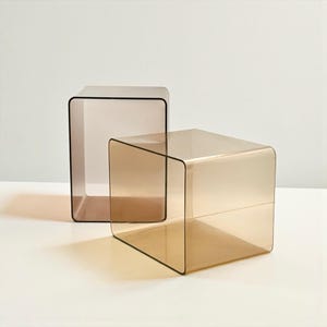 May include: Two modern, transparent acrylic side tables. One is a cube, the other a rectangular shape. Both have rounded corners and a warm, amber-brown hue. The tables are placed on a white surface.