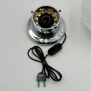 May include: Chrome lamp base with a black electrical cord and plug. The base has a circular opening with a black socket and a white wire.