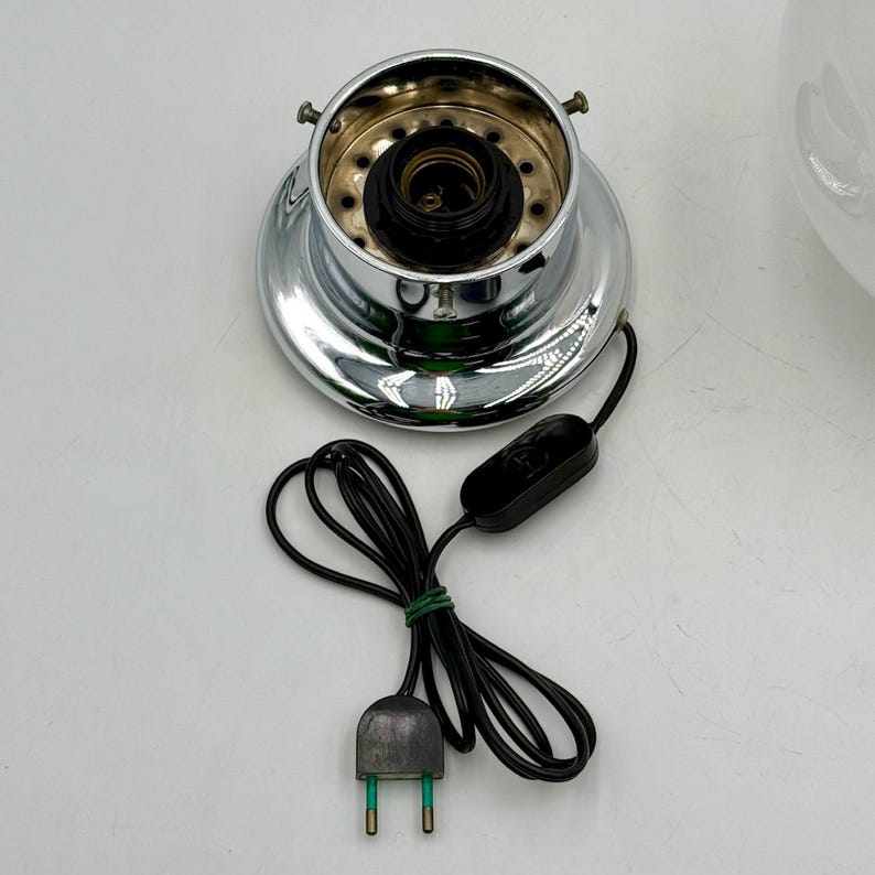 May include: Chrome lamp base with a black electrical cord and plug. The base has a circular opening with a black socket and a white wire.